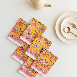 Puede incluir: Seis servilletas amarillas con estampado floral y borde rosa y blanco dispuestas sobre una superficie blanca. También se ven una tetera crema, platos y cubiertos cobrizos. El diseño floral incluye detalles rojos y verdes.