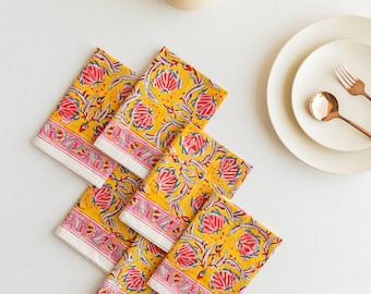 May include: Six yellow floral print napkins with a pink and white border are arranged on a white surface. A cream teapot, plates, and copper-coloured cutlery are also visible. The floral design includes red and green details.