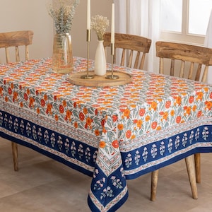 Può includere: Una tovaglia rettangolare con un motivo floreale arancione e bianco, bordata da una fascia blu navy con dettagli floreali. Il tavolo è apparecchiato con sedie in legno, candele e vasi, creando un'atmosfera accogliente.
