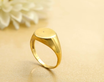 Könnte beinhalten: Ein goldener Siegelring mit glatter, ovaler Oberfläche. Der Ring hat eine polierte Oberfläche und ein klassisches Design. Der Ring wird auf einer hellen Oberfläche mit einer verschwommenen weißen Blume im Hintergrund gezeigt.