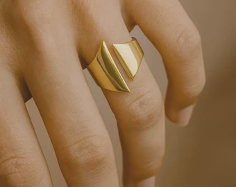 May include: A gold-colored ring with a geometric design. The ring features a split band and a triangular top, creating a modern and minimalist aesthetic. The ring is worn on a finger, showcasing its polished surface.