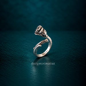 Puede incluir: Un anillo abierto de plata con un capullo de rosa esculpido en un extremo y un diseño de tallo texturizado que se envuelve alrededor del dedo. El metal tiene un acabado oxidado para resaltar los detalles intrincados de los pétalos y las espinas.