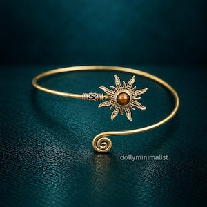 Peut inclure: Un bracelet ouvert doré orné d'un motif en forme de soleil avec une perle brune centrale. Le bracelet possède une tige fine et lisse qui se termine par une spirale à une extrémité et par le motif solaire à l'autre. Le texte dollyminimalist est visible.