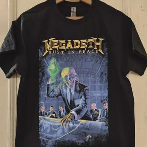 Könnte beinhalten: Schwarzes T-Shirt mit dem Bandnamen "Megadeth" und dem Albumtitel "Rust in Peace". Die Grafik zeigt ein Skelett im Anzug, das einen leuchtend grünen Gegenstand hält, vor futuristischem Hintergrund. Detailliertes Design.
