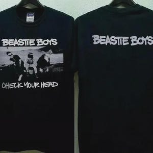 Könnte beinhalten: Schwarzes T-Shirt mit dem weißen Schriftzug "Beastie Boys" über einem Schwarz-Weiß-Bild der Bandmitglieder. Unter dem Bild steht der Text "Check Your Head". Auf der Rückseite des Shirts steht ebenfalls der Bandname in Weiß.