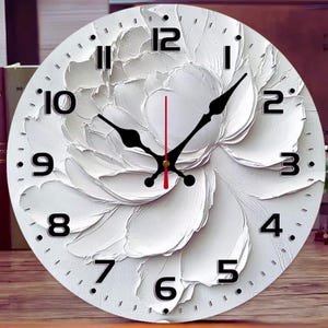 Pode incluir: Relógio de parede redondo branco com um design floral texturizado. O mostrador do relógio tem algarismos e ponteiros pretos, com um ponteiro de segundos vermelho. O design do relógio assemelha-se a uma grande flor branca.