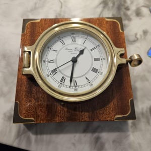 Könnte beinhalten: Eine Quarzuhr im Vintage-Stil mit weißem Zifferblatt und schwarzen römischen Ziffern, eingefasst in einen Messingrahmen. Die Uhr ist auf einem quadratischen Holzsockel mit Messing-Eckakzenten montiert. Das Zifferblatt zeigt "Geneva Hamburg since 1856".