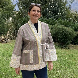 May include: A long-sleeved jacket featuring a muted taupe and grey plaid pattern on the main body, contrasted with floral print cuffs, hem, and pocket trim. The garment includes a bright yellow piped collar edge and a thin white fabric tie closure.
