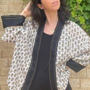 May include: A white open-front jacket featuring a repeating paisley pattern in shades of gold and black. The garment has wide black cuffs and a black front placket, both accented with thin gold zigzag stitching along the edges and hemline.