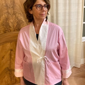 May include: A light pink kimono-style jacket featuring a subtle dark pink polka dot pattern, a white front panel with a delicate texture, long sleeves with wide cuffs, two front pockets, and a thin white ribbon tie closure at the center front.