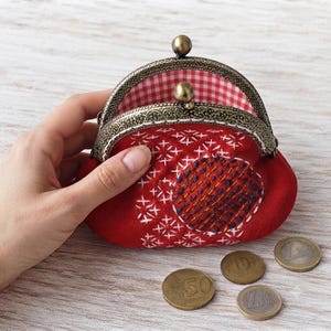 Peut inclure: Un porte-monnaie en tissu rouge avec un fermoir à cliquet en métal bronze. L'extérieur présente des broderies en croix blanches et un empiècement circulaire avec des points bleu foncé et jaunes. L'intérieur est doublé de tissu vichy rouge et blanc.