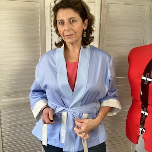 Puede incluir: Chaqueta estilo kimono azul claro con estampado de cuadros blancos y azules. La chaqueta tiene un cinturón a juego y ribetes blancos en las mangas. La modelo lleva la chaqueta sobre un top rojo y vaqueros azul oscuro.