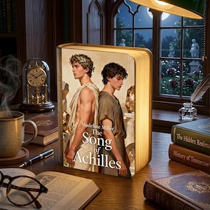 Op de afbeelding: Een boekvormige lamp met de cover van "The Song of Achilles" van Madeline Miller. Op een houten bureau met een kop koffie, boeken en een vintage klok. De cover toont twee mannen in klassieke kleding, verlicht door een warme gloed.