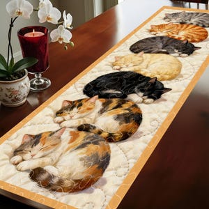 Könnte beinhalten: Rechteckiger Tischläufer mit einem sich wiederholenden Muster von schlafenden Katzen in verschiedenen Farben, darunter Schildpatt, orangefarben getigert und schwarz. Die Katzen sind auf einem cremefarbenen, gesteppten Hintergrund mit einem orangefarbenen Rand angeordnet.