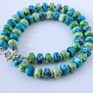 Puede incluir: Collar de cuentas redondas en tonos azules, verdes y amarillos. Las cuentas tienen un aspecto veteado. El collar tiene un cierre plateado y se muestra sobre una superficie blanca. Una joya.