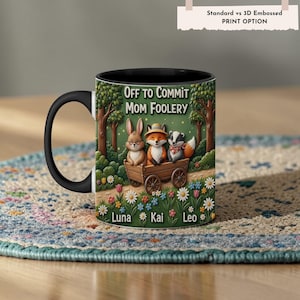 May include: Black ceramic mug with a cartoon design. The mug shows a rabbit, fox, and badger in a wooden cart, with the text "OFF TO COMMIT MOM FOOLERY." The names "Luna," "Kai," and "Leo" are also displayed. The mug has a black handle.