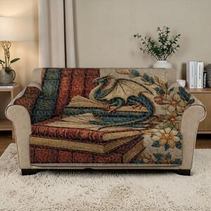 May include: A sofa cover featuring a fantasy design. The cover depicts a blue and teal dragon resting on a stack of books. The books are in shades of red, brown, and gold. Floral accents are also present.