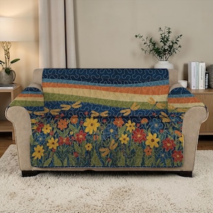 Op de afbeelding: Een bankhoes met een bloemen- en libellenontwerp. De hoes heeft een blauwe achtergrond met kleurrijke bloemen, libellen en een streepjespatroon in tinten oranje, groen en geel. De hoes is over een beige bank gedrapeerd.