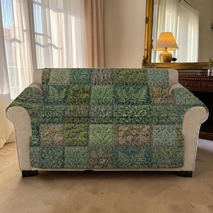 Può includere: Un divano con una coperta trapuntata verde e marrone a tema botanico. La coperta presenta un design patchwork con vari motivi di piante e foglie. Il divano ha una struttura di colore neutro ed è posto davanti a una finestra.