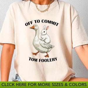 May include: Cream-colored t-shirt featuring a cartoon goose carrying a white rabbit. The text "OFF TO COMMIT TOM FOOLERY" is printed above and below the illustration in black. The bottom of the shirt has a green banner with white text.