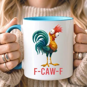 May include: White ceramic mug with a blue interior and handle. The mug features a cartoon rooster illustration with red, yellow, green, and blue colors. The text "F-CAW-F" is printed in red below the rooster.