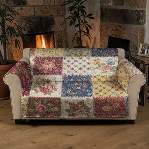 May include: A beige sofa with a patchwork cover featuring floral patterns in red, blue, yellow, and cream. The cover is placed over the sofa's seat and armrests. The background includes a fireplace and potted plants.