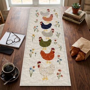 May include: A cream-coloured quilted table runner with a repeating pattern of chickens in various colours, including grey, orange, and green, and floral accents. The runner is displayed on a wooden table.
