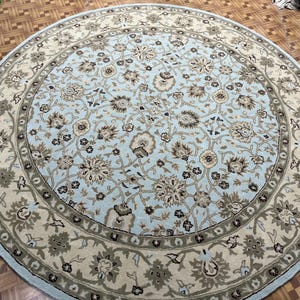 May include: A round area rug with a light blue center featuring a floral pattern in shades of brown, beige, and gray. The rug has a beige border with a gray floral design and a light blue edge.