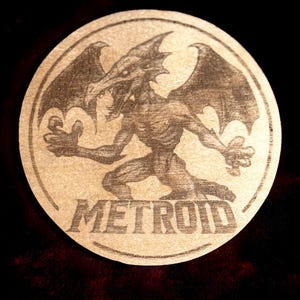 K&ouml;nnte beinhalten: Ein runder, beigefarbener Untersetzer mit einer detaillierten Illustration einer drachenartigen Kreatur mit gro&szlig;en Fl&uuml;geln und dem Wort "METROID" in fetten Buchstaben. Das Design ist vor einem dunklen Hintergrund.