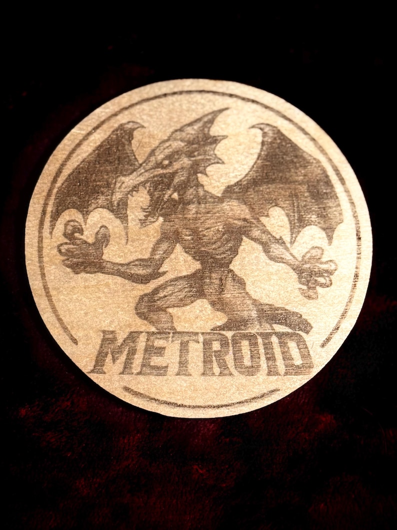 K&ouml;nnte beinhalten: Ein runder, beigefarbener Untersetzer mit einer detaillierten Illustration einer drachenartigen Kreatur mit gro&szlig;en Fl&uuml;geln und dem Wort "METROID" in fetten Buchstaben. Das Design ist vor einem dunklen Hintergrund.