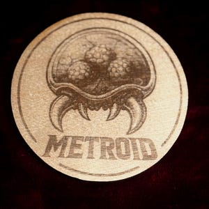 K&ouml;nnte beinhalten: Ein runder, hellbrauner Untersetzer mit einer detaillierten Gravur eines Metroid-Kreatur. Das Wort "METROID" ist darunter gedruckt. Das Bild hat einen dunklen Hintergrund, mit einer detaillierten Illustration.