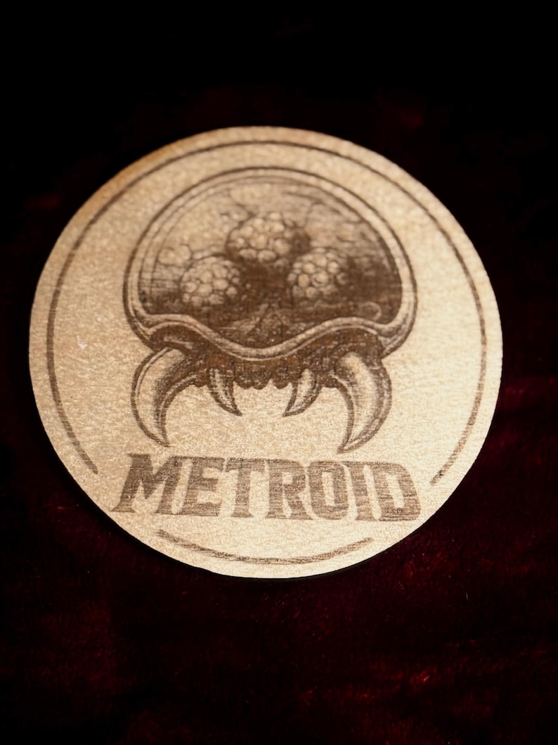 K&ouml;nnte beinhalten: Ein runder, hellbrauner Untersetzer mit einer detaillierten Gravur eines Metroid-Kreatur. Das Wort "METROID" ist darunter gedruckt. Das Bild hat einen dunklen Hintergrund, mit einer detaillierten Illustration.