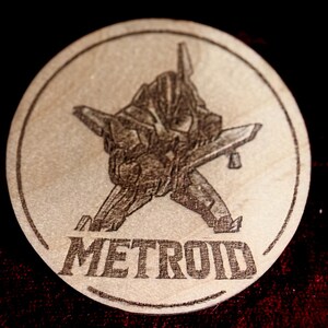 K&ouml;nnte beinhalten: Ein runder Holzuntersetzer mit einem lasergravierten Design. Das Design zeigt eine detaillierte Illustration eines futuristischen Raumschiffs und das Wort "METROID". Der Untersetzer ist hellbraun.