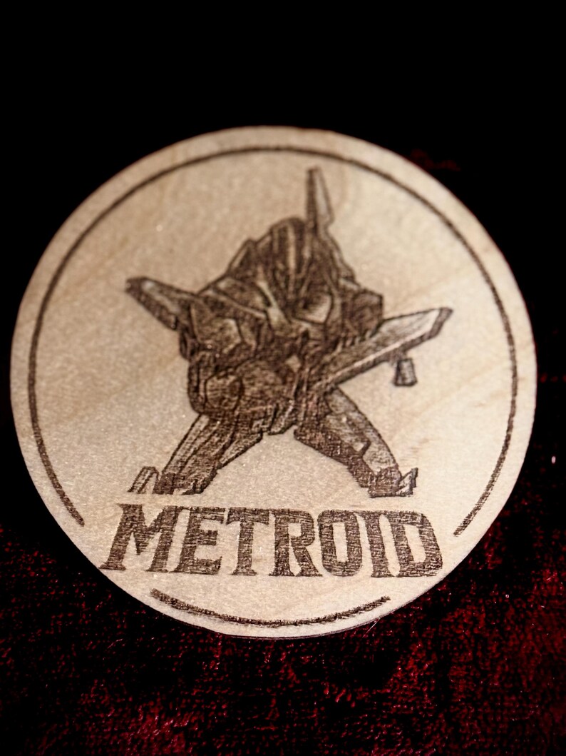 K&ouml;nnte beinhalten: Ein runder Holzuntersetzer mit einem lasergravierten Design. Das Design zeigt eine detaillierte Illustration eines futuristischen Raumschiffs und das Wort "METROID". Der Untersetzer ist hellbraun.