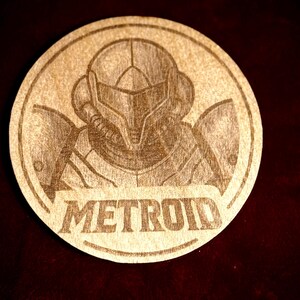 K&ouml;nnte beinhalten: Ein runder Holzuntersetzer mit einer detaillierten Gravur eines Charakters aus der Videospielserie Metroid. Das Wort "METROID" ist darunter eingraviert. Der Untersetzer ist aus hellem Holz gefertigt.