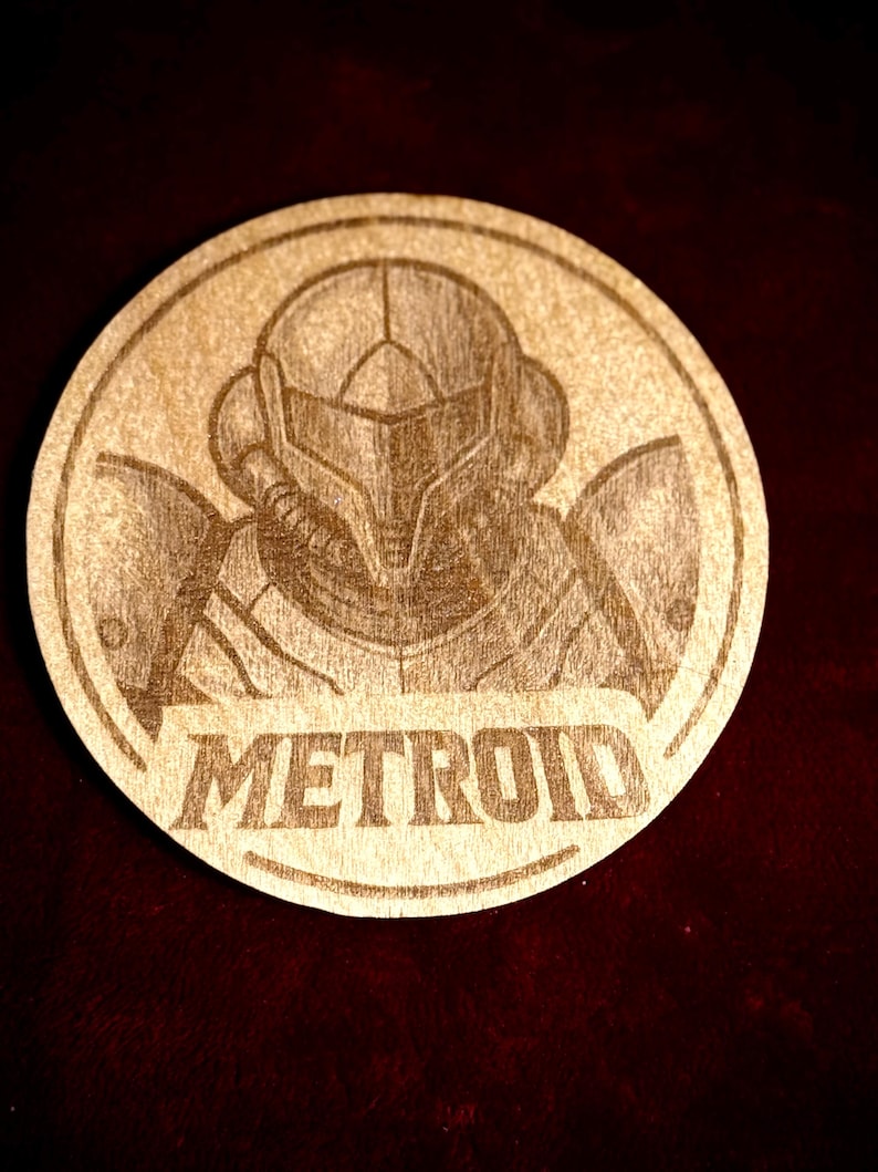 K&ouml;nnte beinhalten: Ein runder Holzuntersetzer mit einer detaillierten Gravur eines Charakters aus der Videospielserie Metroid. Das Wort "METROID" ist darunter eingraviert. Der Untersetzer ist aus hellem Holz gefertigt.