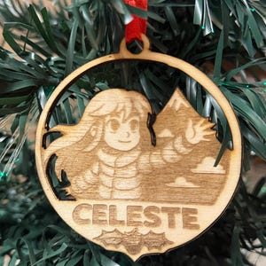 May include: Wooden Christmas ornament featuring a laser-cut design of a character and mountain scene. The word "CELESTE" is engraved at the bottom. The ornament hangs from a red ribbon and is displayed on a Christmas tree.