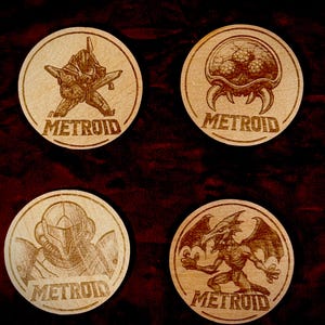 K&ouml;nnte beinhalten: Vier Holzuntersetzer mit lasergravierten Metroid-Designs. Jeder Untersetzer zeigt einen anderen Charakter oder eine Kreatur aus dem Spiel, mit dem Wort "METROID" darunter. Die Untersetzer sind kreisf&ouml;rmig und aus hellem Holz gefertigt.