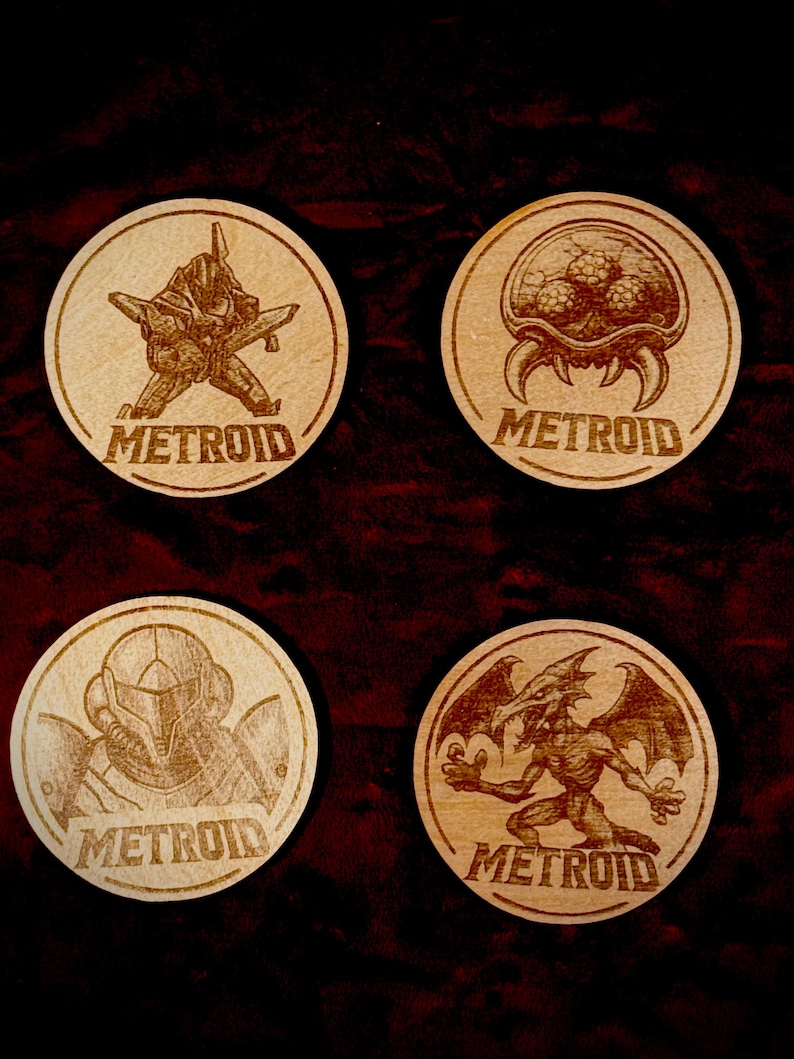 K&ouml;nnte beinhalten: Vier Holzuntersetzer mit lasergravierten Metroid-Designs. Jeder Untersetzer zeigt einen anderen Charakter oder eine Kreatur aus dem Spiel, mit dem Wort "METROID" darunter. Die Untersetzer sind kreisf&ouml;rmig und aus hellem Holz gefertigt.