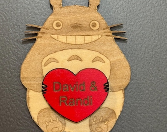Könnte beinhalten: Holzdekoration in Totoro-Form, die ein rotes Herz hält. Der Totoro ist hellbraun mit einem lächelnden Gesicht und Schnurrhaaren. Das rote Herz trägt die Namen "David & Randi". Der Hintergrund ist einfarbig grau.