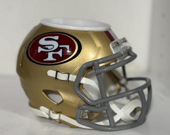 May include: A miniature gold and red San Francisco 49ers football helmet. The helmet features the team's logo on the side and a grey facemask. The helmet is a decorative item.