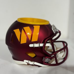 May include: A burgundy football helmet desk accessory featuring a prominent yellow letter W logo on the side. The helmet has a yellow interior rim, a burgundy face mask, and white chin straps. The top of the helmet is open, creating a hollow container.