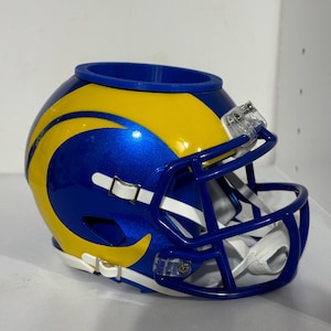 Könnte beinhalten: Ein Miniatur-Footballhelm in Blau und Gelb, mit einem blauen Gesichtsschutz und einem weißen Kinnriemen. Der Helm hat eine glänzende Oberfläche und eine kreisförmige Öffnung oben. Das Design umfasst einen gelben Streifen an der Seite.