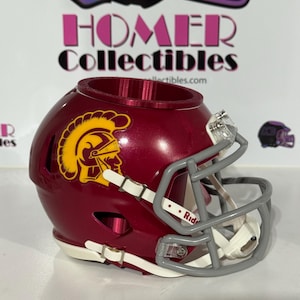 May include: A maroon football helmet planter featuring a yellow Trojan logo on the side. The helmet has a grey face mask, white chin straps, and a hollow top opening. The text "Homer Collectibles" is visible in the background behind the helmet.