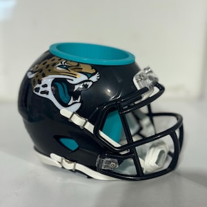May include: A black and teal football helmet-shaped container with a Jacksonville Jaguars logo. The helmet has a black face mask and white accents. The container has a teal interior and a teal rim.