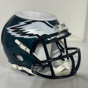 Könnte beinhalten: Ein dunkelgrüner Footballhelm-Wachswärmer mit einem weißen Adlerflügel-Logo an der Seite. Der Helm hat einen schwarzen Gesichtsschutz, einen weißen Kinnriemen und ein weißes, kreisförmiges Becken für Wachs an der Oberseite. Unten ist das Logo Homer Collectibles.