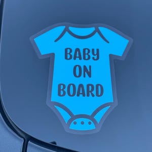 May include: A blue baby onesie-shaped car sticker with the words "BABY ON BOARD" in black. The sticker has a white outline and is adhered to a car window. The design is simple and clear, indicating a baby is in the vehicle.