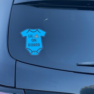 May include: A blue onesie-shaped car sticker with the text "Lil ON BOARD" in white. A cartoon poop emoji is above the word "BOARD". The sticker is affixed to the rear window of a dark gray car.