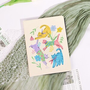 May include: A cream-coloured notebook with a vibrant illustration of cats in various poses and colours, including yellow, blue, pink, and purple, with floral accents. The notebook is set against a backdrop of green fabric.