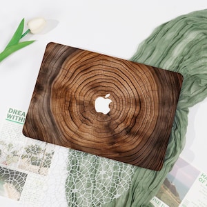 May include: A laptop case featuring a wood grain design with a white Apple logo. The case has a natural, rustic appearance with concentric rings, and is set against a white background with a green fabric.